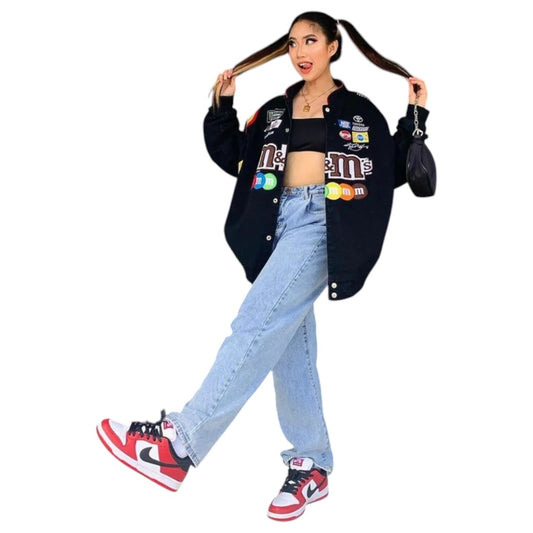 Person wearing a black jacket with colorful patches, black crop top, light blue jeans, and red sneakers on a white background.